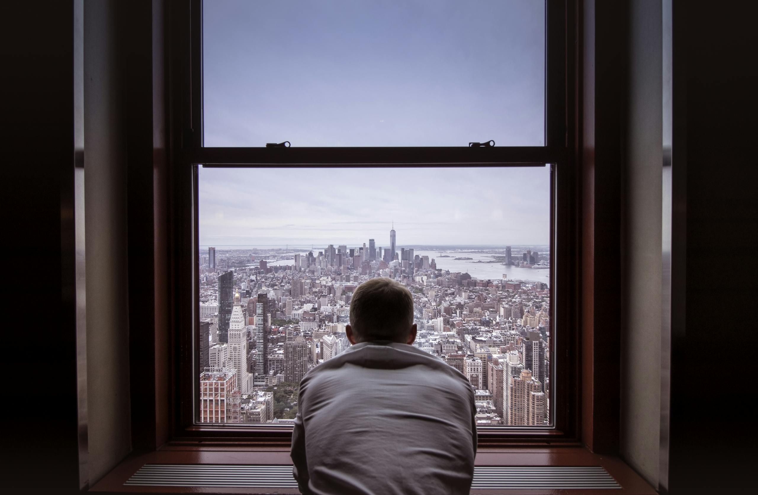 A man reflects while overlooking the iconic New York City skyline through a large window.